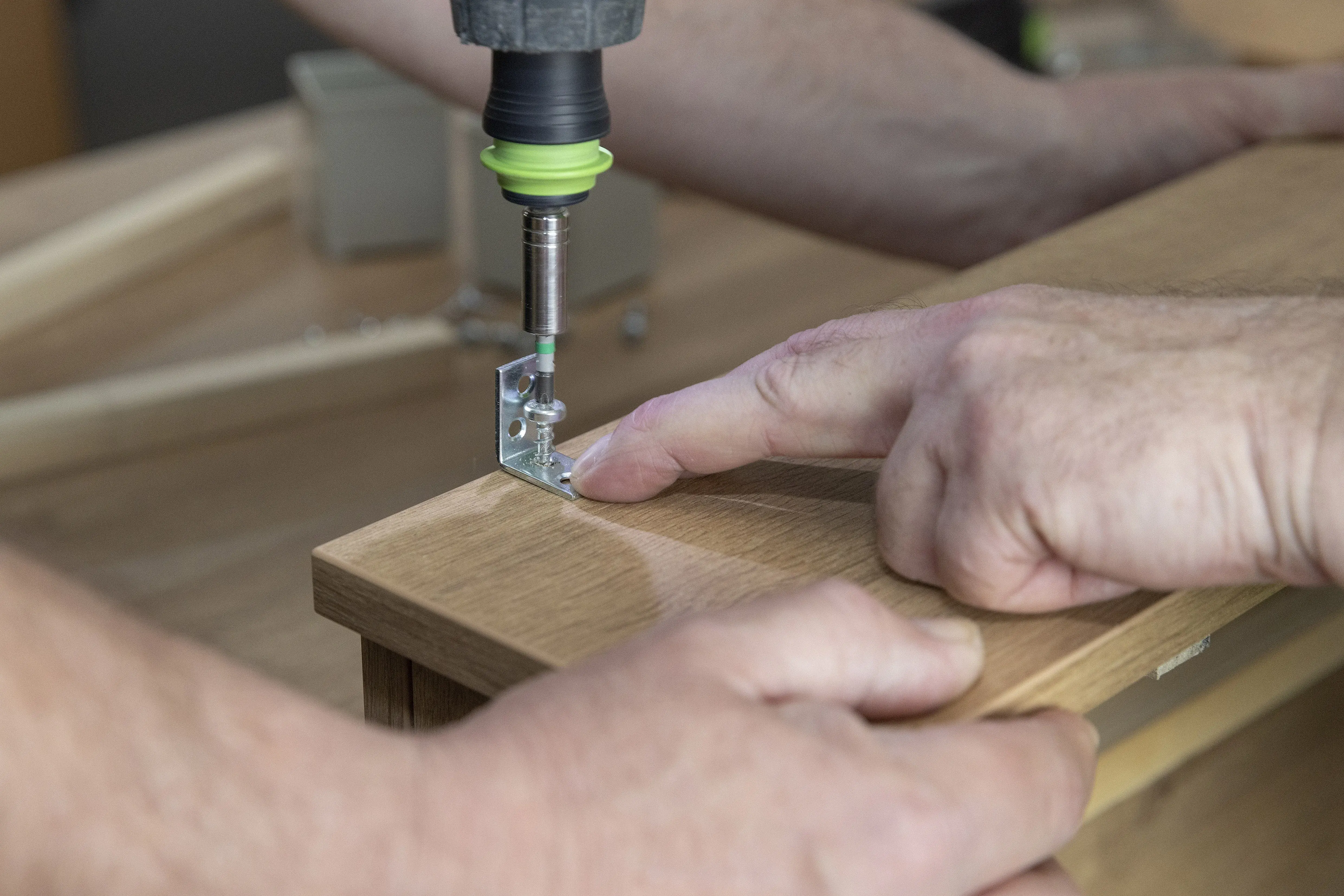 Jemand schraubt ein Metallwinkel mit einer Bohrmaschine an ein Holzbrett.